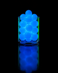 A cluster of glowing blue gel balls inside a clear glass tumbler, centered on a black reflective surface with a blue reflection below.